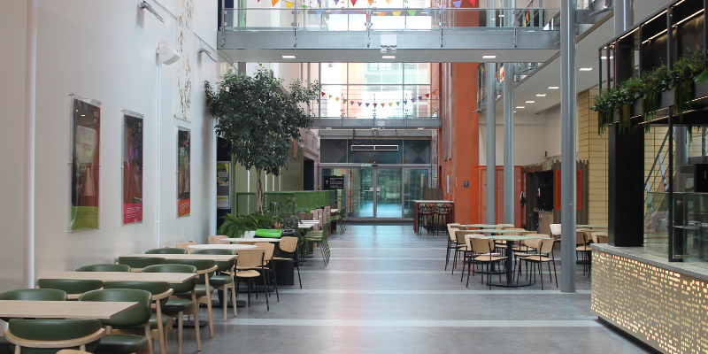 Interior view of The Atrium cafe featuring seating with wooden tables and green chairs along a wide hallway, potted plants, high ceilings, and a serving counter on the right.
